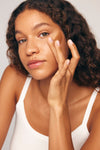 A young woman with curly brown hair gently touches her face, showcasing radiant skin enhanced by Liquid Bronzer v2. Wearing a white tank top, she gazes calmly into the camera against a neutral background.