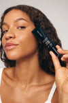 A woman with curly hair and glowing skin holds a black tube of Liquid Bronzer v2 by SALT & STONE near her face, smiling softly. She wears a white top and poses against a neutral background.