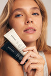 A woman with long blonde hair holds two SALTYFACE Sunscreen & Bronze tubes—one white and one black—close to her face, looking into the camera with a neutral expression.