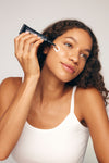 A woman with curly hair in a white tank top applies Sunscreen & Bronze SPF 30 to her cheek from a black tube, smiling gently and looking upward.
