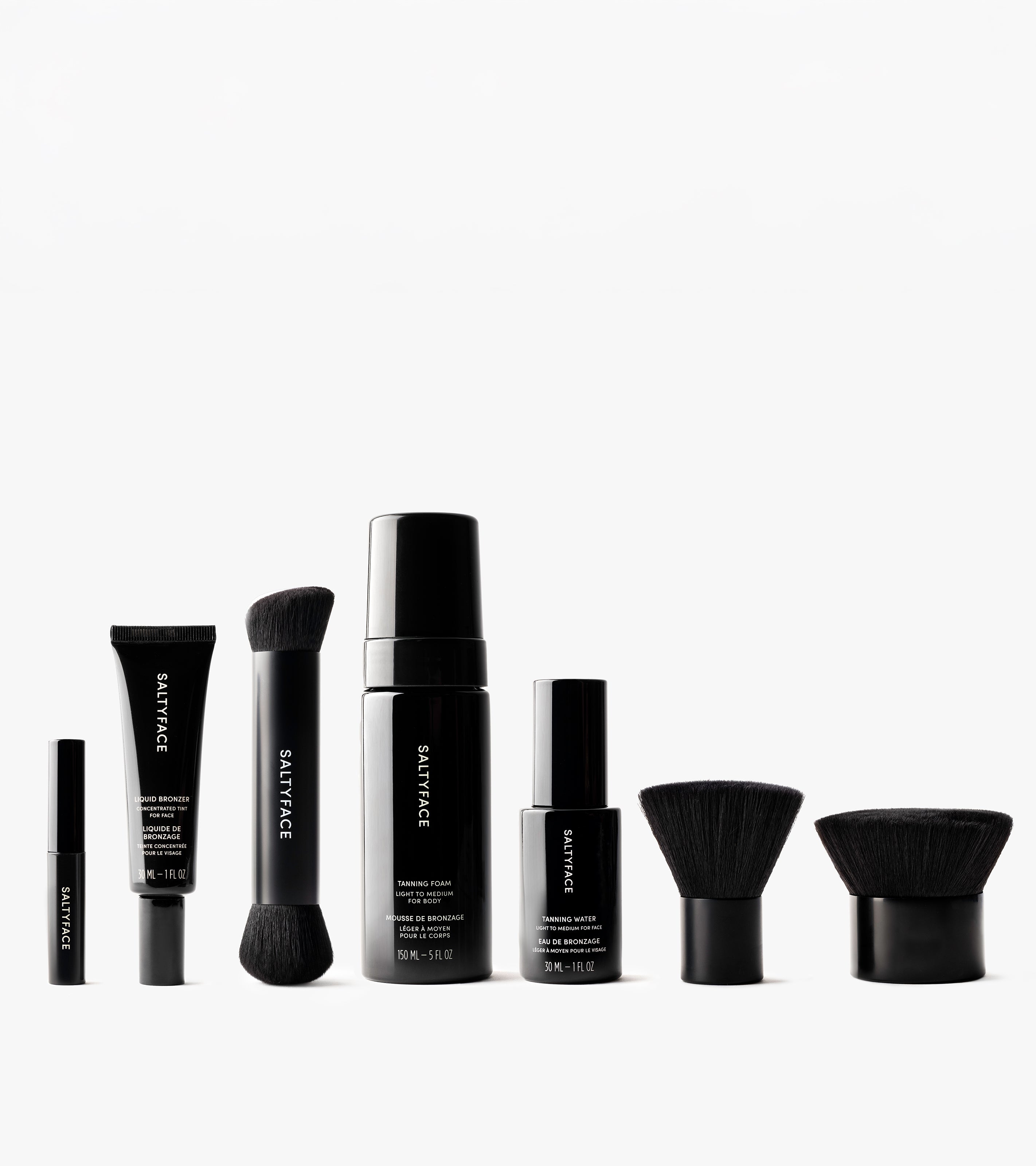 The Saltyface Set features six black makeup products and brushes—tubes, bottles, and two kabuki-style brushes—arranged in a row on white, ideal for creating a natural sun-kissed or bronzed look.