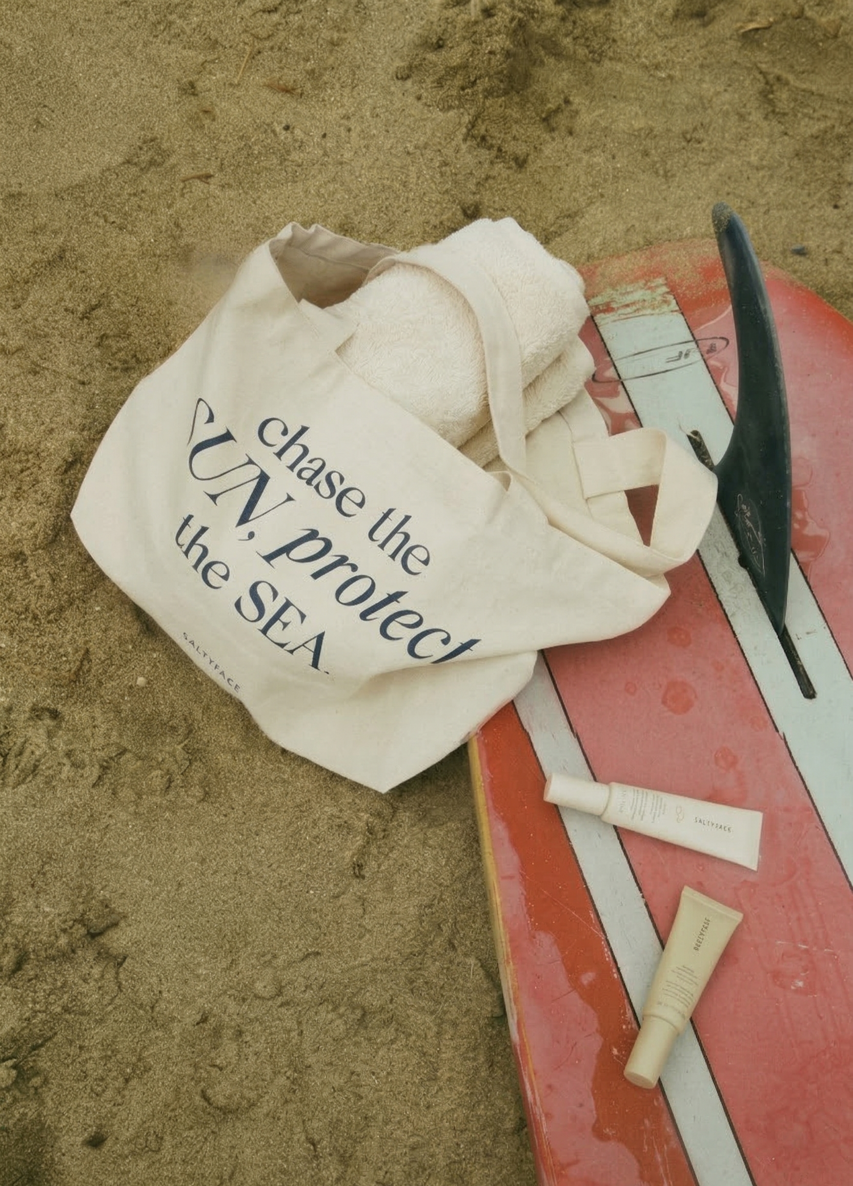 The Tote, a comfy canvas shoulder bag with "chase the UN.protect the SEA," sits on sand by a red surfboard. The tote holds a white towel and two tubes of skincare products rest on the board nearby.