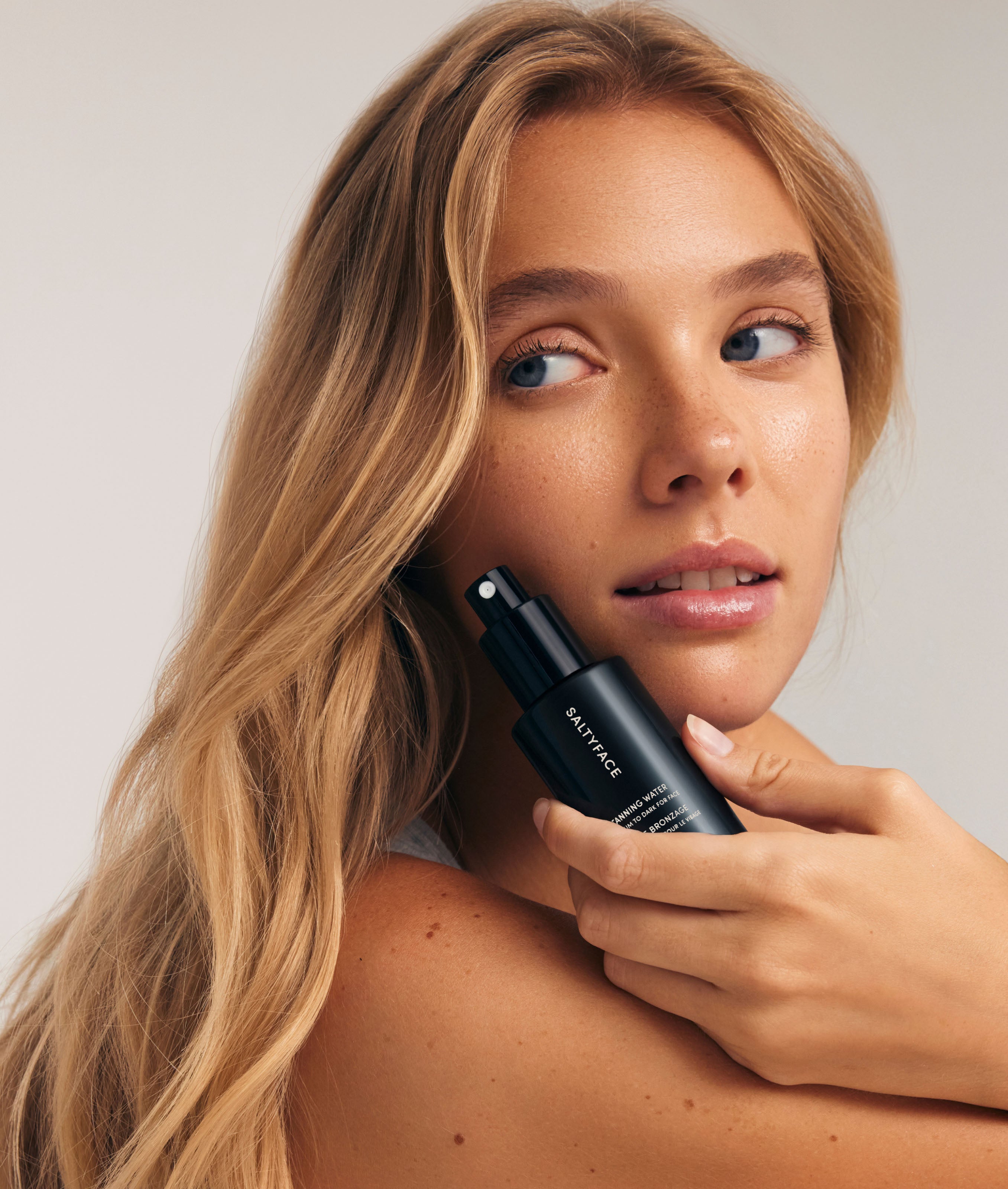 A woman with long blonde hair holds The Complete Face & Body Set near her face, gazing softly to the side against a neutral background.