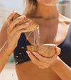 A woman in a bikini pours coconut water between coconut shells, highlighting The Complete Face & Body Set for a flawless tan. The words AVAILABLE IN COCONUT appear in the upper right corner.