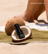 A black bottle of The Face Set sits inside a halved coconut on a beach towel, with sand, a whole coconut, and someone's legs in the background. Text reads AVAILABLE IN COCONUT.
