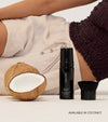 A person in light clothing lies next to half a coconut, a black cosmetic bottle, and a body brush. Text reads: The Body Set—AVAILABLE IN COCONUT for the perfect sun-kissed tan.
