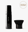 A black bottle labeled The Body Set stands next to a sleek body brush. Above them, a circle says Available in Coconut on a white background, ready to give you a flawless sun-kissed tan.