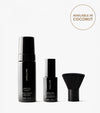 The Essential Face & Body Set, comprising a Tanning Foam, Tanning Water, and face brush in sleek black packaging, stands upright on a white background. A round label highlights its Coconut scent option.