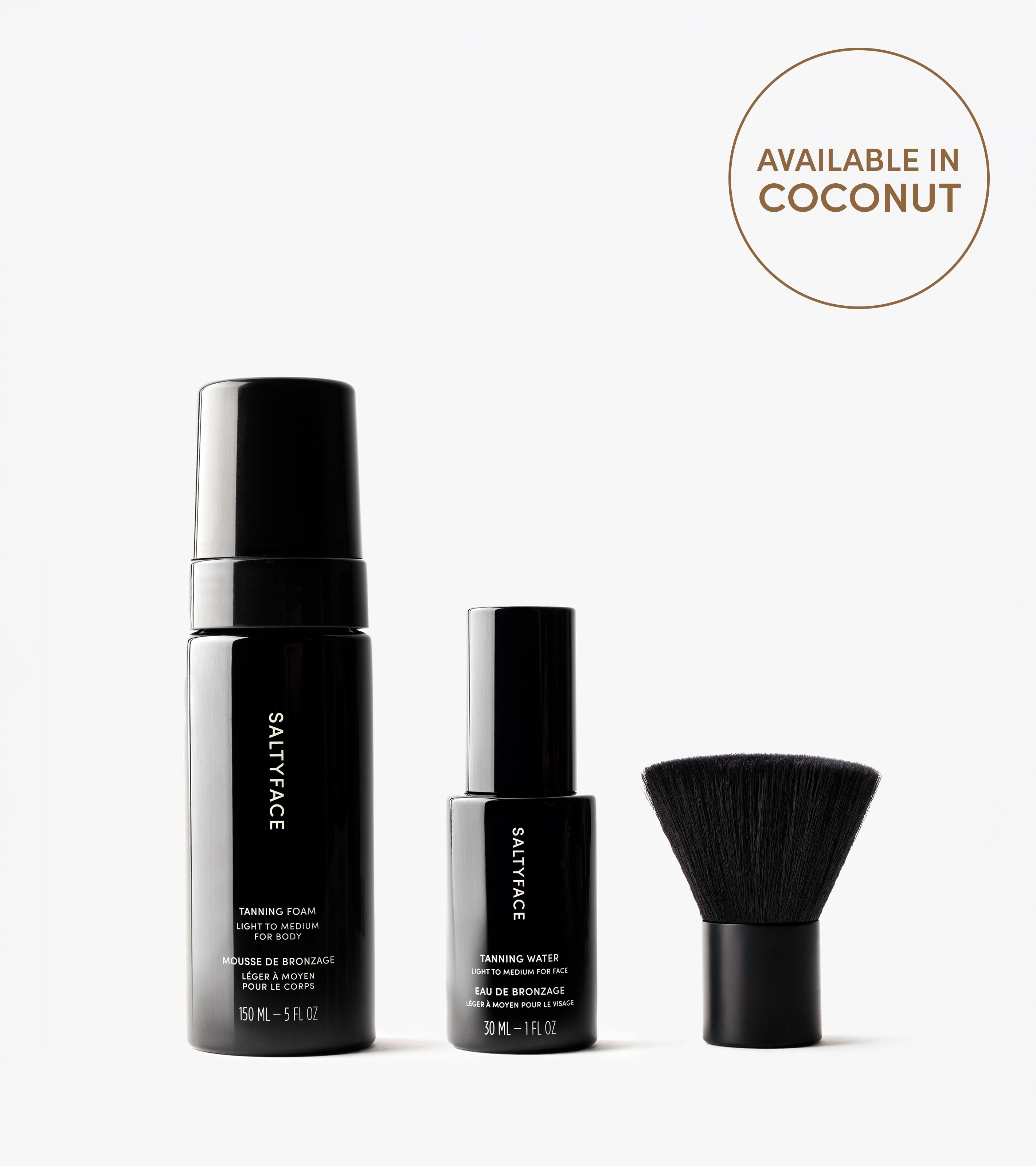 The Essential Face & Body Set, comprising a Tanning Foam, Tanning Water, and face brush in sleek black packaging, stands upright on a white background. A round label highlights its Coconut scent option.