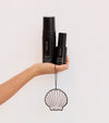 A hand holds The Coconut Set—two black skincare bottles—against a plain light background, with a decorative seashell-shaped ornament hanging by a string below.