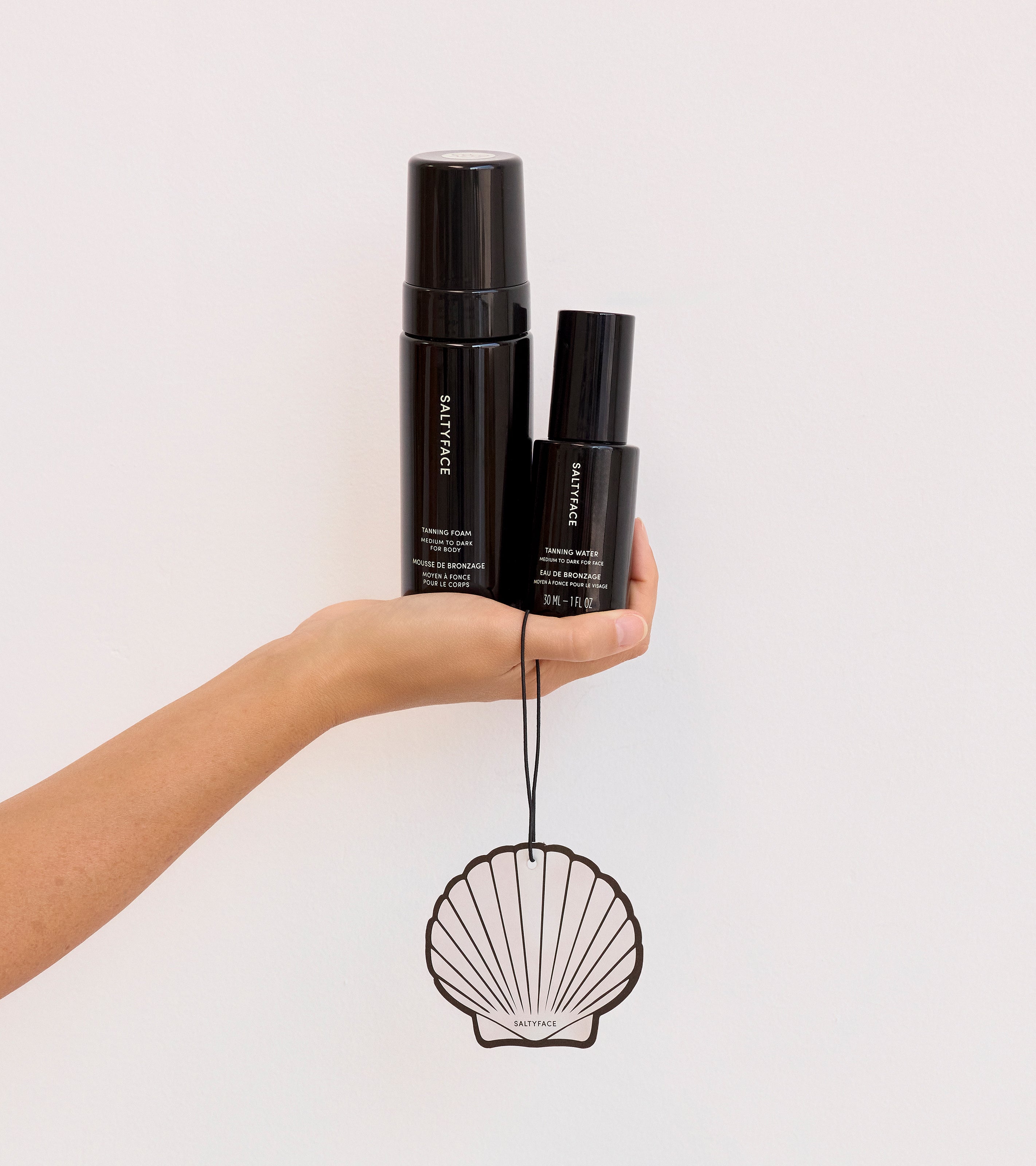 A hand holds The Coconut Set—two black skincare bottles—against a plain light background, with a decorative seashell-shaped ornament hanging by a string below.