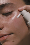 A close-up of a person with eyes closed as they apply Daily Sun Shield mineral sunscreen SPF 30 to their cheek using a pump dispenser. Their skin looks dewy and well-moisturized.