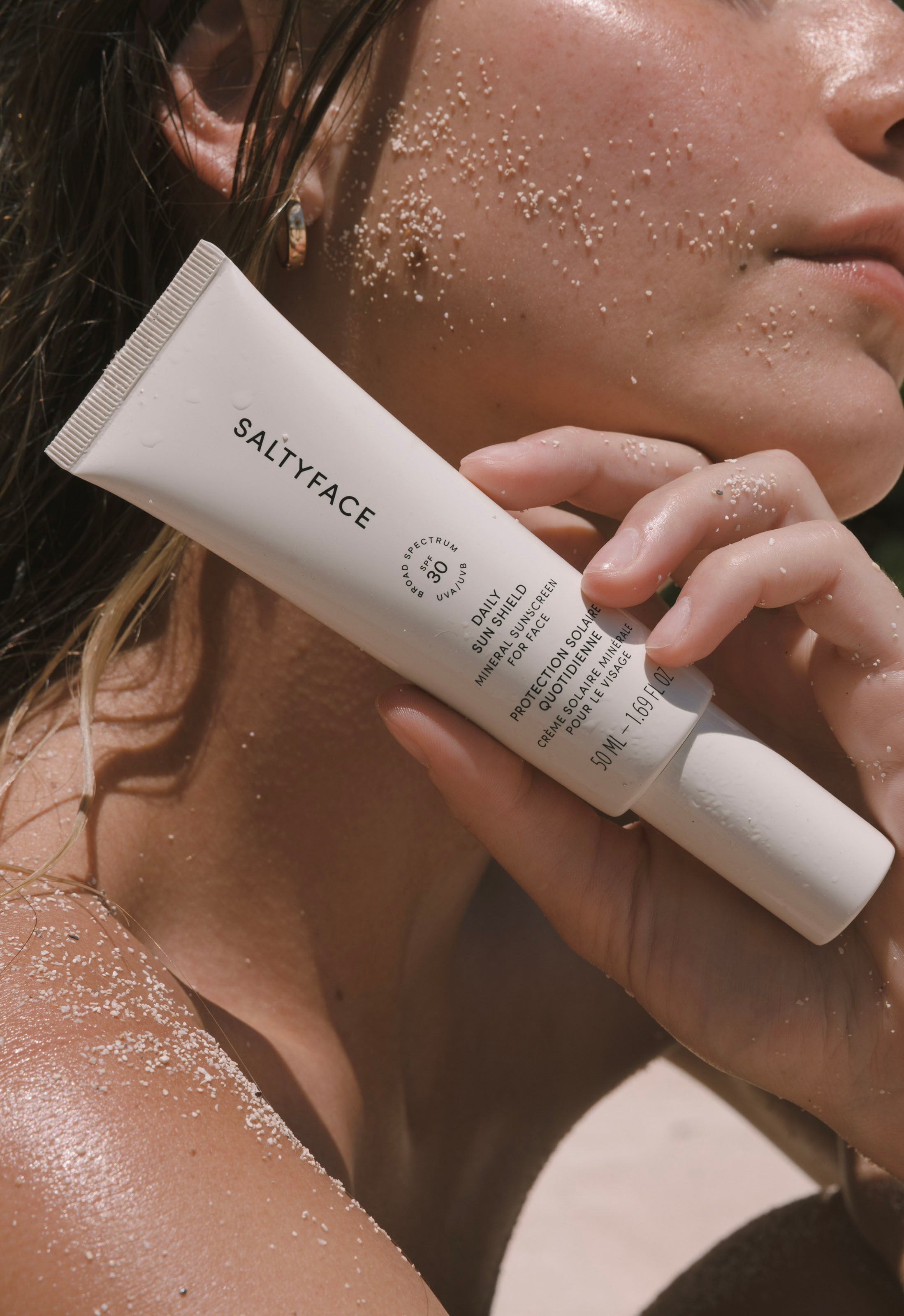 A woman with sand on her face and shoulder holds a tube of Daily Sun Shield SPF 30 mineral sunscreen near her face while basking in sunlight.