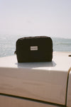 A black The Everywhere Bag sits on the white hood of a vehicle, serving as a sleek travel pouch with the ocean and hazy sky in the background.