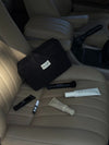 The Everywhere Bag, a black zippered pouch, sits on the beige leather passenger seat of a car alongside assorted skincare and makeup products near the gear shift.