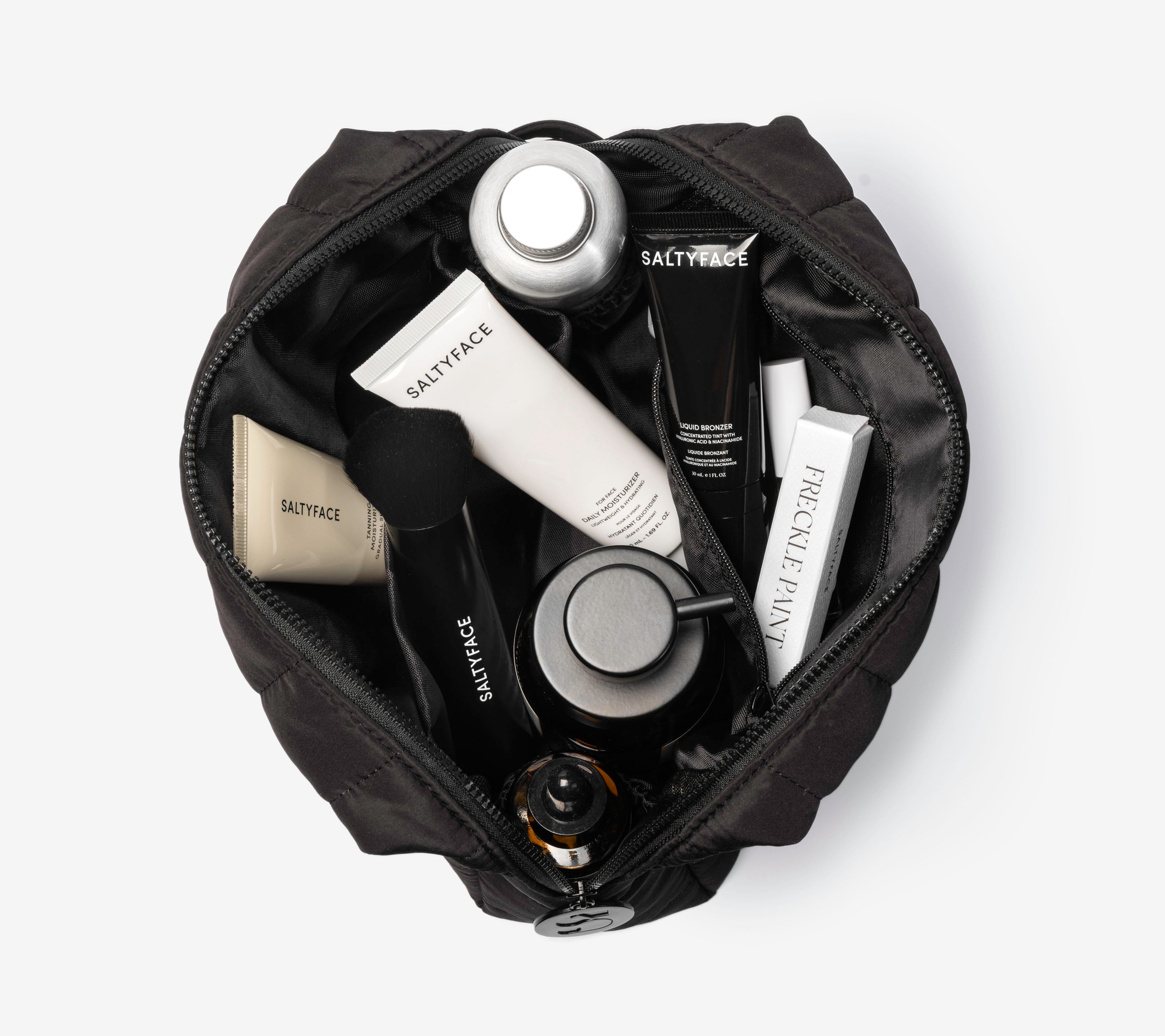 The Everywhere Bag, a black zippered pouch, opens to display neatly organized skincare products—tubes, bottles, a brush—and items labeled Saltyface and Furcile Faynt, making it the perfect travel companion against a white background.
