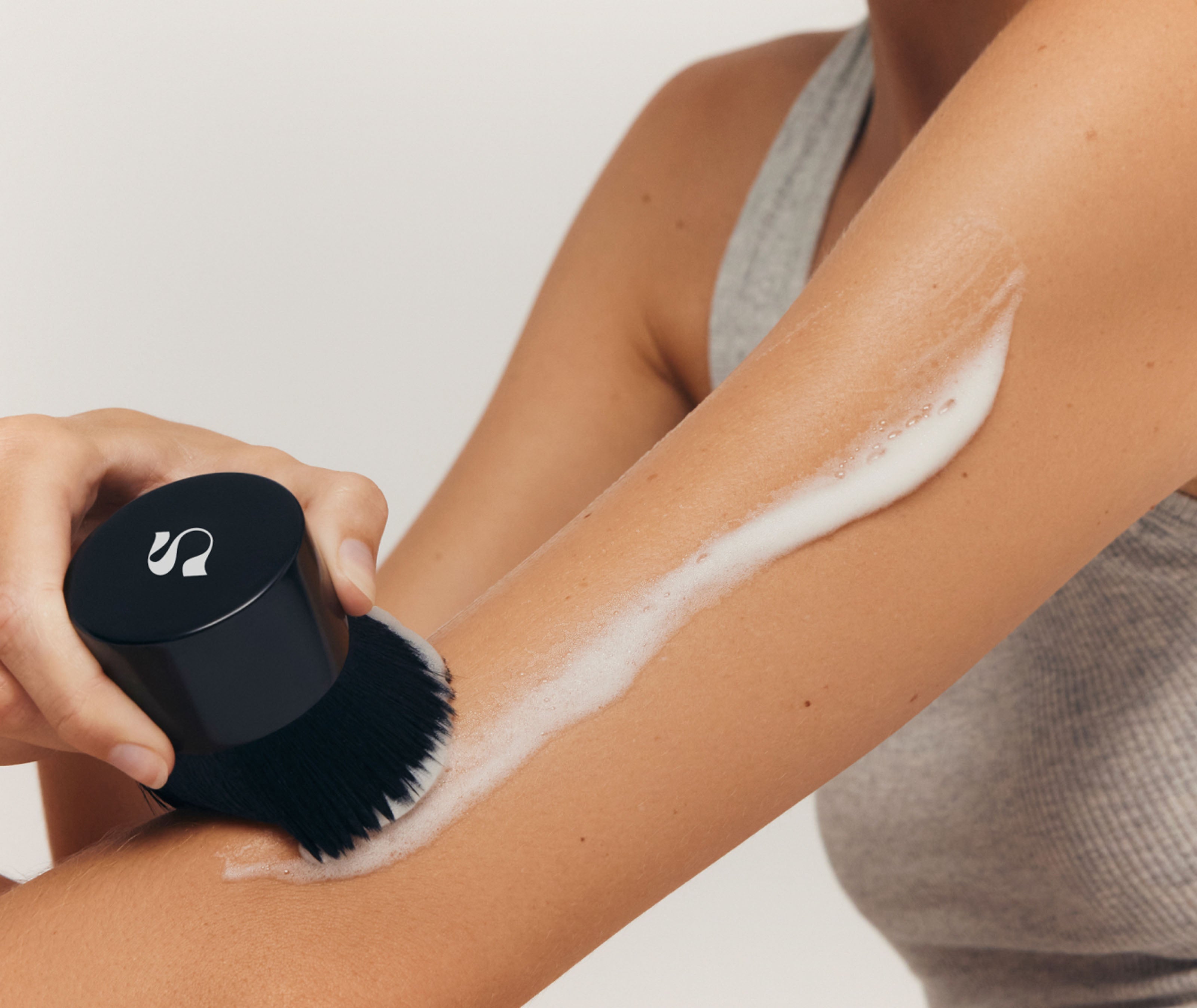 Person using a black brush with bristles to apply tanning foam to their arm against a neutral background.