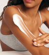 A woman in a gray bra gently applies Tanning Foam, a water-to-foam formula, to her upper arm with her fingers. Only her lower face and torso are shown.