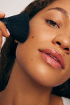 A close-up of a person using The Essential Face & Body Set to apply makeup with a large brush, highlighting smooth, glowing skin, natural lips, and brown eyes.