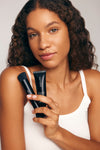 A woman with curly hair in a white tank top smiles slightly while holding The Saltyface Set—a makeup brush and foundation tube—against a neutral background, ready for a natural look.