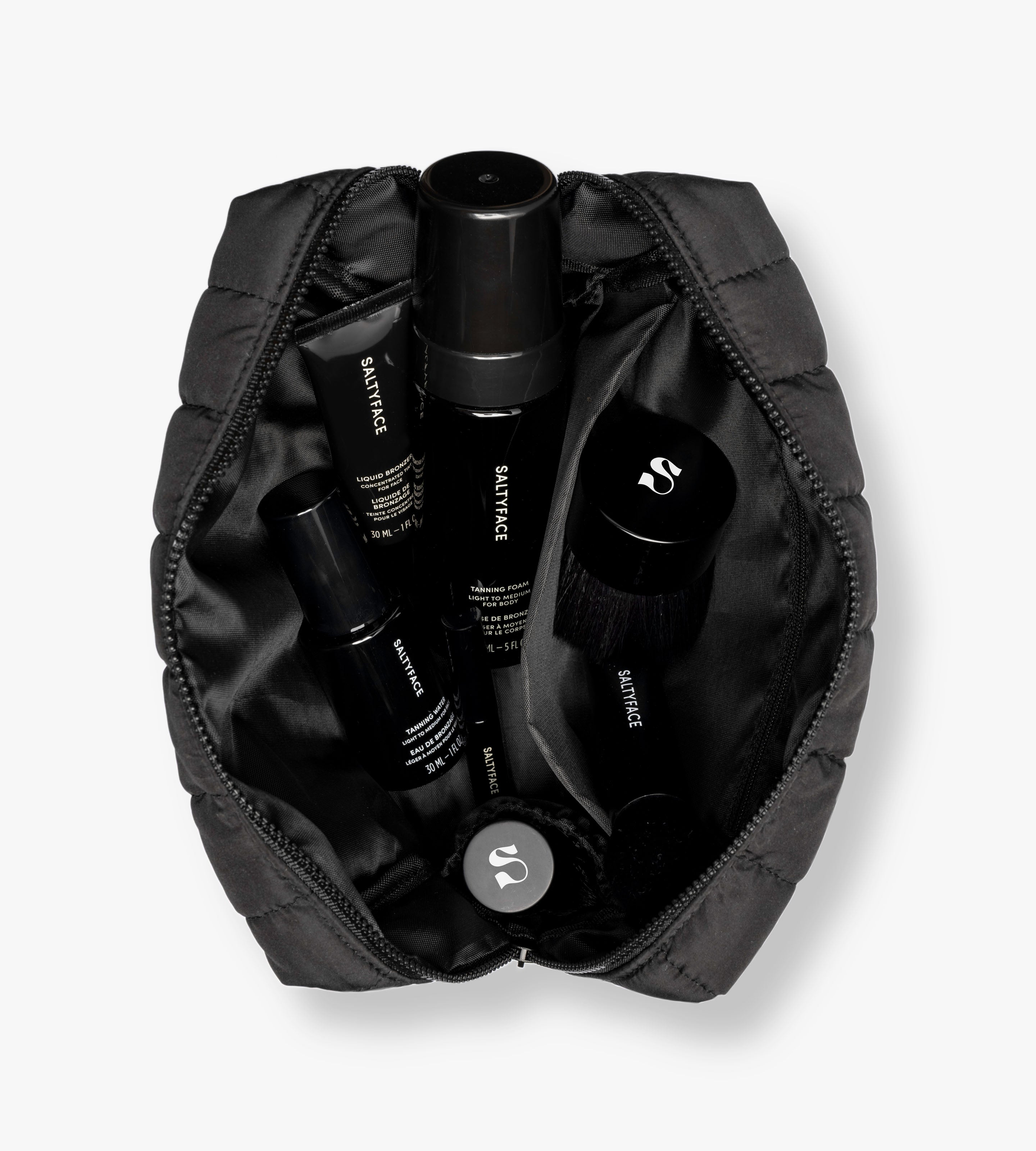 The Saltyface Set features a black zip pouch holding best-selling face and body care bottles and a round brush, all labeled in white text, arranged for an effortless, natural sun-kissed glow.