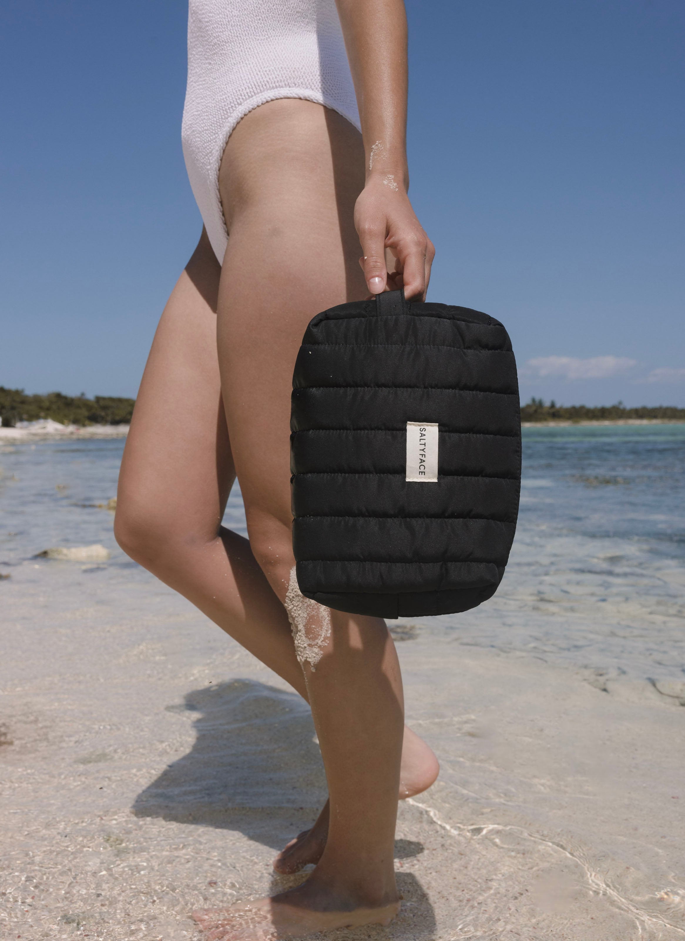 A person in a white swimsuit stands on a sandy beach near the water, holding The Everywhere Bag, a small black quilted travel bag. Blue sky and distant trees are visible in the background.